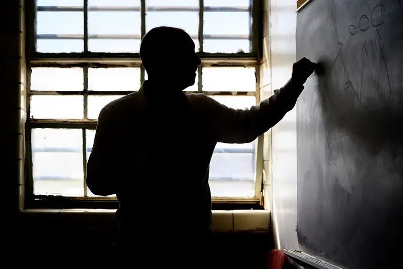 A man stands in profile writing on a chalkboard. He is silhouetted against a bright window.